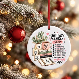 May include: A round white ceramic ornament with a red ribbon featuring an illustration of a potted plant, a candle, a stack of books, and an open notebook. The text reads Positivity is a choice, I choose, and lists six positive affirmations.