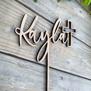 K&ouml;nnte beinhalten: Holz-Kuchentopper mit dem Namen "Kaylan" in Kursivschrift und einem Holzkreuz.