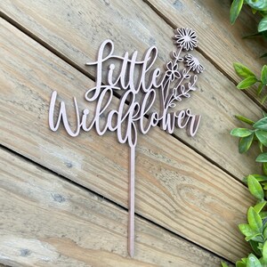 Puede incluir: Topper de pastel de madera con el texto "Little Wildflower" y un grupo de flores estilizadas.