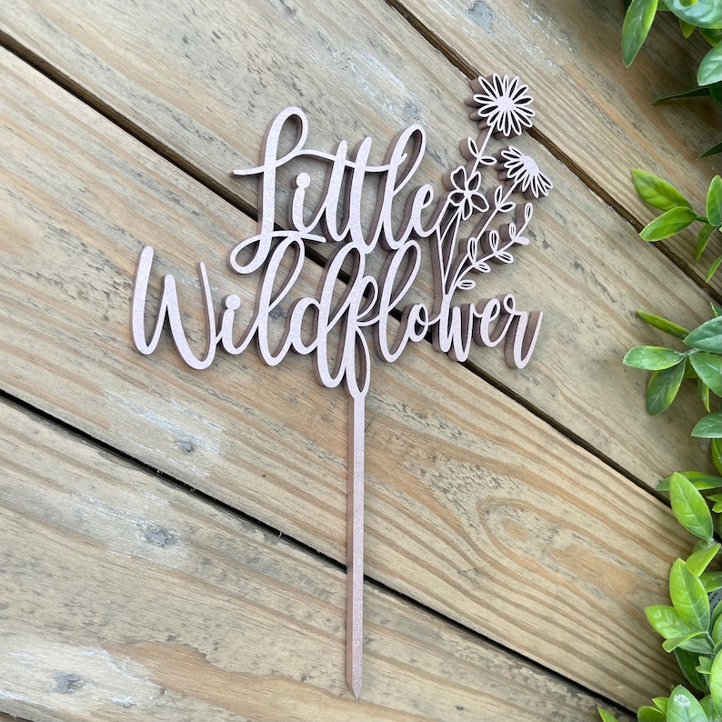Puede incluir: Topper de pastel de madera con el texto "Little Wildflower" y un grupo de flores estilizadas.