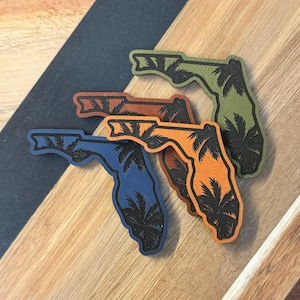 May include: Four leather Florida state outline stickers with palm trees in different colors: blue, orange, brown, and green.