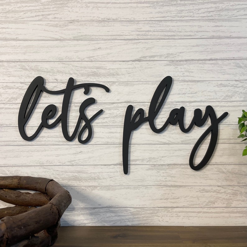 K&ouml;nnte beinhalten: Schwarzes Holzschild mit den Worten "let's play" in kursivschrift. Das Schild ist an einer wei&szlig;en Holzplankenwand befestigt.