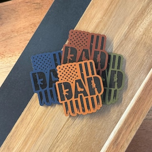 Op de afbeelding: Vier rechthoekige patches met een vlagontwerp en het woord "DAD" in zwart. De patches zijn blauw, oranje, bruin en groen. Het vlagontwerp bevat sterren en verticale strepen, met een geweersilhouet.