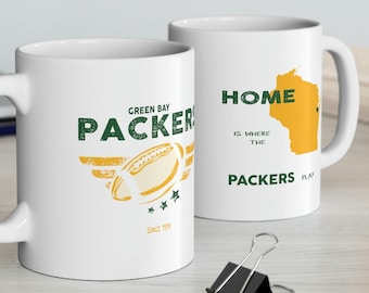 May include: Two white ceramic mugs with green and yellow designs. The mug on the left has the text "Green Bay Packers" and a football with wings. The mug on the right has the text "Home is where the Packers play" and an outline of the state of Wisconsin.