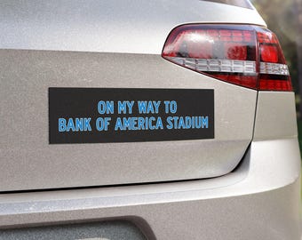 Op de afbeelding: Een zwarte bumper sticker met blauwe tekst die "On my way to Bank of America Stadium" zegt. De sticker is bevestigd aan de achterkant van een zilveren auto.