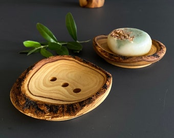 May include: Two wooden soap dishes made from olive wood. The dishes are round and have a natural, rustic look. One dish has a bar of soap with green and gold flecks on top.