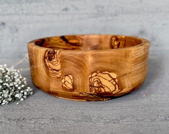 May include: A small, round wooden bowl made from olive wood. The bowl has a natural wood finish and a unique grain pattern.