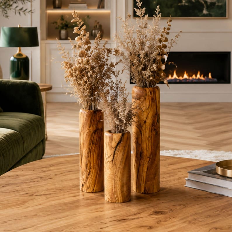 May include: Three wooden vases of varying heights, filled with dried ornamental grasses, sit on a round wooden table. The vases have a natural wood grain pattern. The background includes a fireplace, a green velvet sofa, and a decorative lamp.