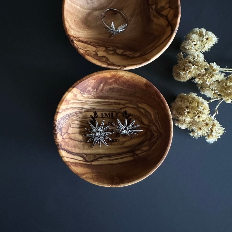 May include: Two wooden bowls with a natural wood grain finish. The smaller bowl has the text "EMILY & 2023" engraved on the inside. Both bowls contain silver jewelry, including a ring and earrings.