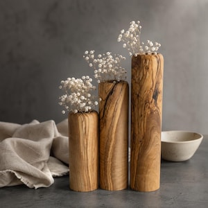 May include: Three wooden vases of varying heights, each filled with delicate white flowers. The vases have a natural wood grain finish, showcasing shades of brown and tan. The arrangement is set against a neutral backdrop, with a linen cloth and a ceramic bowl in the background.