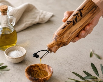 May include: A cylindrical olive wood oil dispenser bottle featuring a dark engraved olive branch design. The bottle has a stainless steel pour spout with a black cap, currently pouring golden olive oil into a matching small olive wood bowl.