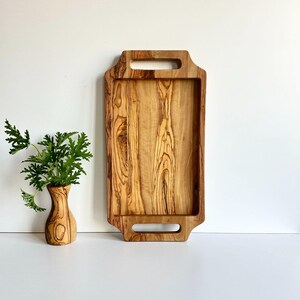 May include: A rectangular wooden serving tray with two handles. The tray is made of light brown wood with dark brown streaks. The tray is sitting on a white surface.
