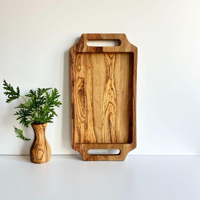 May include: A rectangular wooden serving tray with two handles. The tray is made of light brown wood with dark brown streaks. The tray is sitting on a white surface.
