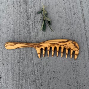 May include: A wooden comb with a natural wood grain finish. The comb has a long handle and a wide tooth design.