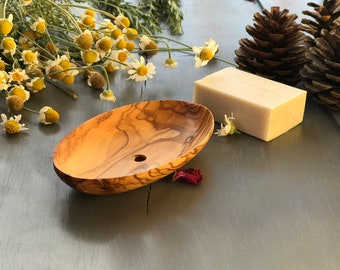 May include: A wooden soap dish made from olive wood with a drainage hole. The dish is oval shaped and has a natural wood grain. The dish is sitting on a gray wooden surface.