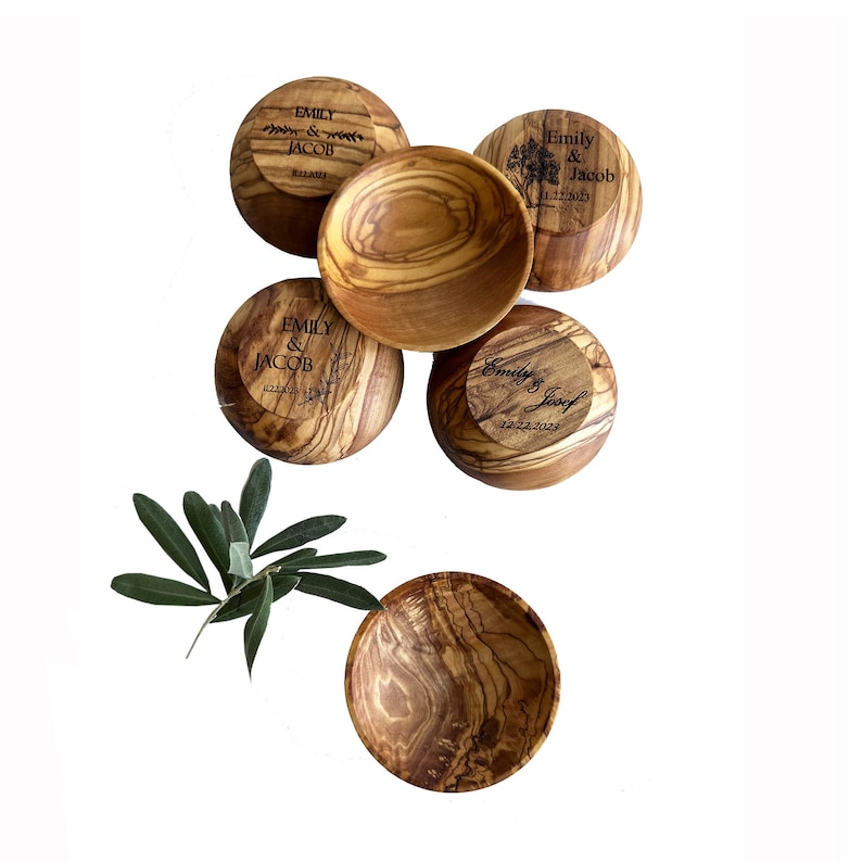May include: Five small olive wood bowls with engraved names and dates. The bowls are arranged in a circle with a sprig of greenery in the center.