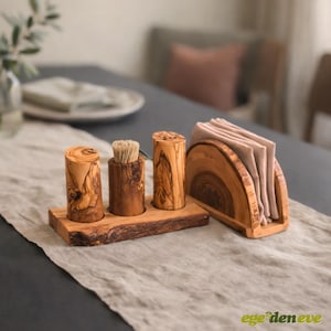 Puede incluir: Un juego de accesorios de cocina de madera de olivo que incluye una base rectangular con tres soportes cilíndricos para sal, pimienta y palillos, junto a un servilletero semicircular a juego con varias servilletas de tela beige plegadas.
