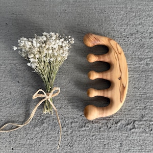 Può includere: Un pettine di legno con manico curvo e denti arrotondati, accanto a un piccolo bouquet di fiori bianchi secchi legati con spago. Il pettine è in legno chiaro con venature visibili.