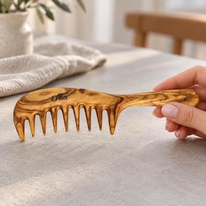 May include: A wide-tooth hair comb crafted from light brown wood with a distinct dark grain pattern. The comb features a smooth, curved handle and the name Alice engraved in a black script font on the upper portion of the comb body.