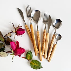 Può includere: Un set di sei posate d'argento con manici in legno. Le posate includono forchette, cucchiai e un coltello. I manici sono di colore marrone chiaro con venature marroni scure.