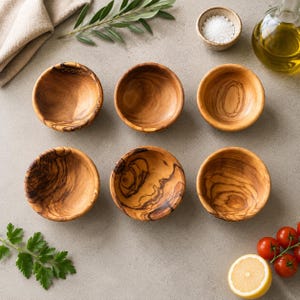 Può includere: Un set di sei piccole ciotole rotonde realizzate in legno d'ulivo naturale, ognuna caratterizzata da un motivo a venature unico con tonalità marroni e beige. Le ciotole sono disposte in due file da tre, evidenziando la superficie liscia e levigata.