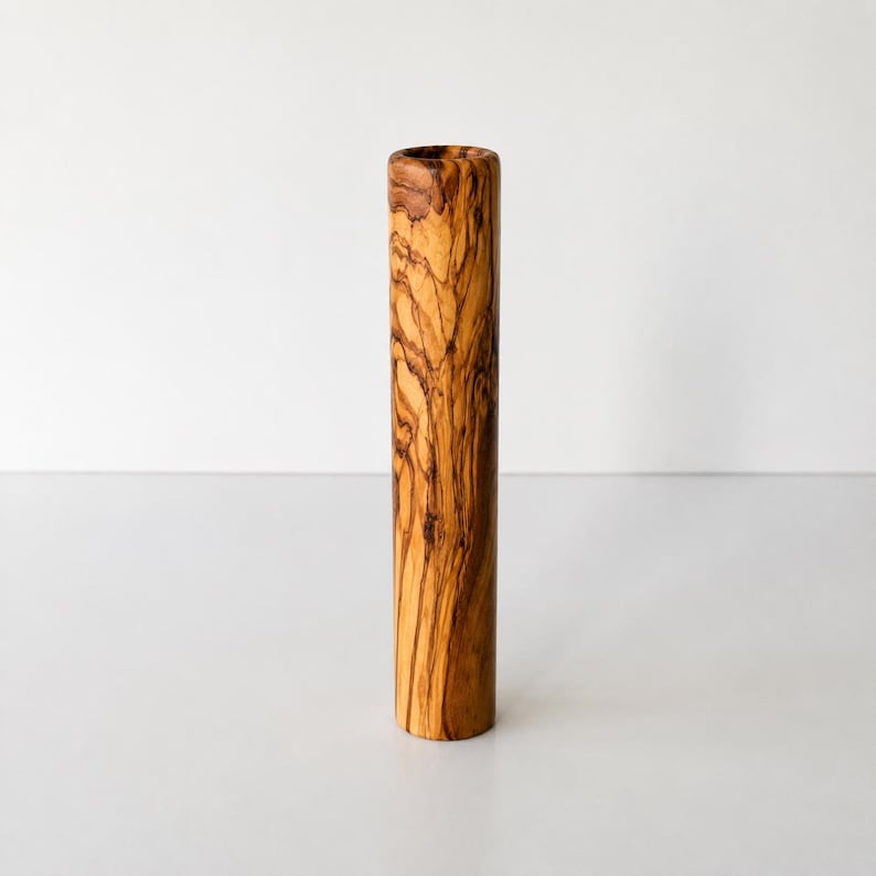 May include: A cylindrical vase made of olive wood. The vase has a natural wood grain pattern in shades of brown and tan. The vase is standing upright against a white background.