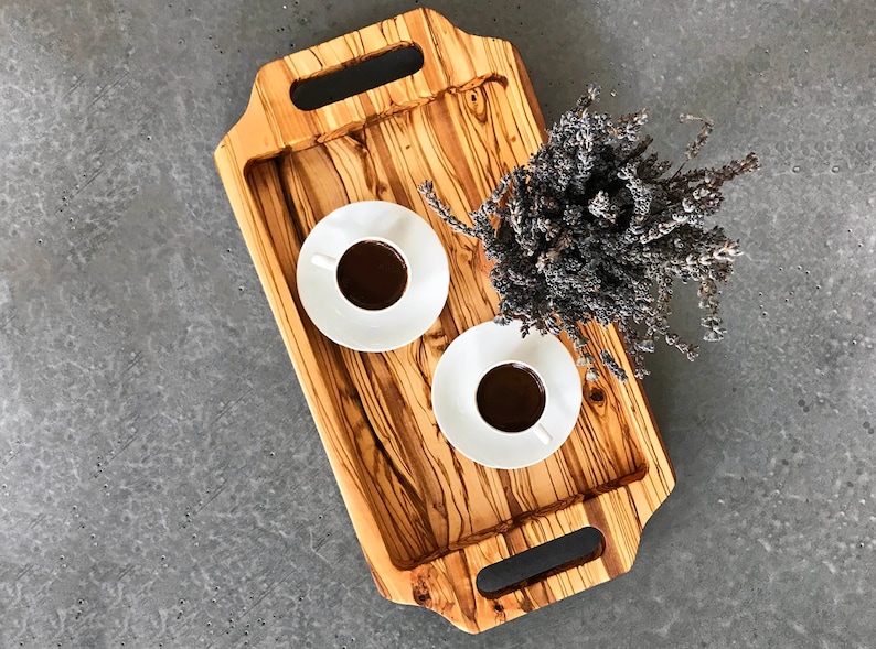 May include: A wooden serving tray with two handles on each side. The tray is made of light brown wood with dark brown streaks. There are two white ceramic cups with coffee on the tray. A bouquet of dried lavender is on the tray.