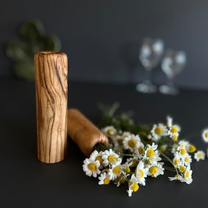 May include: Two wooden bottle stoppers with a natural wood grain pattern. One stopper stands upright, the other lies horizontally next to a bouquet of white daisies with yellow centers. The background is dark, with two wine glasses visible.