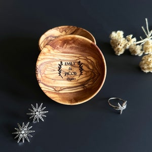 May include: Two small, round, wooden bowls with a natural wood grain pattern. The bowl on the right has the names "Emily & Jacob" and the date "12.22.2023" engraved on it. The bowls are on a black surface with a silver ring and two silver star-shaped earrings.