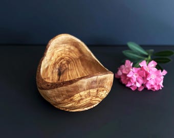 May include: A handcrafted wooden bowl with a unique, organic shape. The bowl is made from olive wood, showcasing a rich, warm brown color with intricate grain patterns. A cluster of pink flowers and green leaves are next to the bowl.