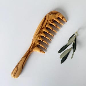 Könnte beinhalten: Ein Holzkamm mit natürlicher Holzmaserung. Der Kamm hat einen langen Griff und einen breiten, gebogenen Kopf mit 10 Zähnen. Der Kamm ist aus Olivenholz.