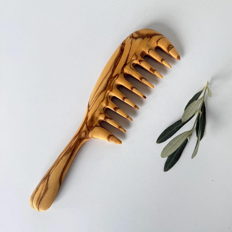 May include: A wooden comb with a natural wood grain pattern. The comb has a long handle and a wide, curved head with 10 teeth. The comb is made of olive wood.