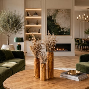 May include: A luxurious living room with a green velvet sofa and armchair. A round wooden coffee table holds books and a brass bowl. Three wooden vases with dried flowers sit in the center. A fireplace and artwork are in the background.