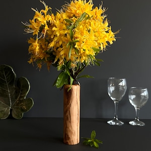 May include: A wooden vase holds a vibrant bouquet of yellow flowers with green leaves. The vase has a cylindrical shape and a natural wood grain finish. Two wine glasses are in the background, suggesting a decorative floral arrangement.