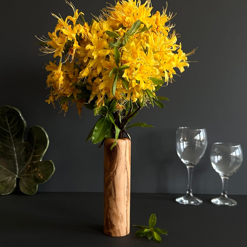 May include: A wooden vase holds a vibrant bouquet of yellow flowers with green leaves. The vase has a cylindrical shape and a natural wood grain finish. Two wine glasses are in the background, suggesting a decorative floral arrangement.