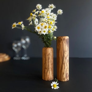 May include: Two wooden vases with a natural wood grain pattern. One vase holds a bouquet of white and yellow daisies. The other vase is empty. A single daisy rests on the surface. The vases are cylindrical and have a rustic aesthetic.