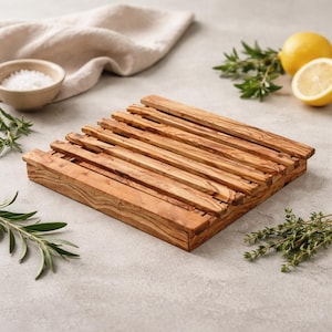 May include: A square wooden trivet made of light brown wood with a slatted design. The trivet is surrounded by fresh herbs, a bowl of salt, and a lemon. The trivet is designed to protect surfaces from hot cookware.