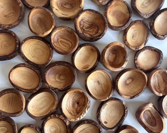 May include: A collection of small, irregular wooden bowls crafted from olive wood, featuring natural light brown wood grain patterns and dark, textured bark edges. Each bowl has a unique, organic shape with a smooth, polished interior surface.