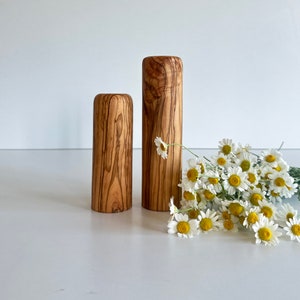 May include: Two wooden cylindrical vases with a natural wood grain pattern. The vases are different heights, with one taller than the other. A bouquet of white daisies with yellow centers rests beside the vases on a white surface.
