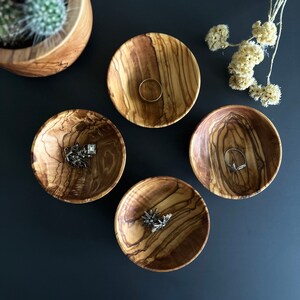 May include: Four small wooden bowls with natural wood grain patterns. Each bowl has a different design and is filled with jewelry, including rings and a chain.