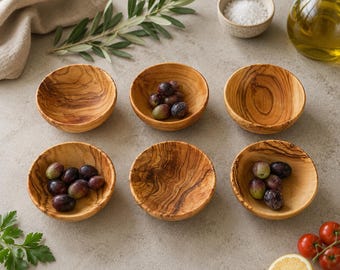 May include: A set of six small, round bowls crafted from olive wood, each featuring a distinct natural grain pattern. Three of the bowls are empty, while the other three contain small, dark purple olives.