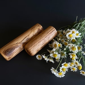 May include: Two wooden rolling pins with a natural wood grain pattern are displayed on a dark surface, accompanied by a bouquet of white daisies with yellow centers. The rolling pins are cylindrical.