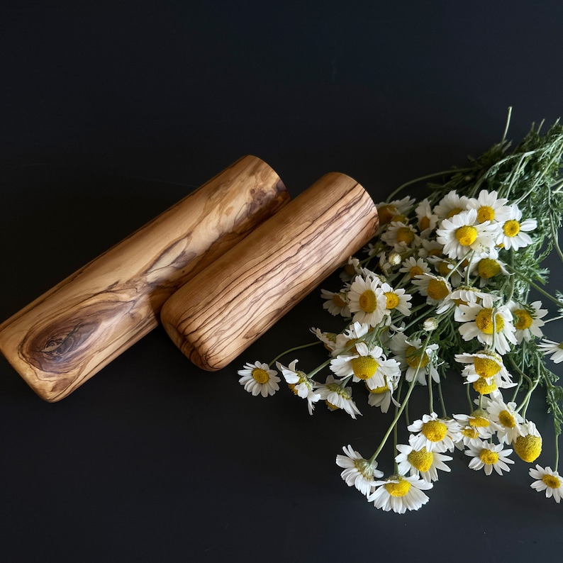 May include: Two wooden rolling pins with a natural wood grain pattern are displayed on a dark surface, accompanied by a bouquet of white daisies with yellow centers. The rolling pins are cylindrical.