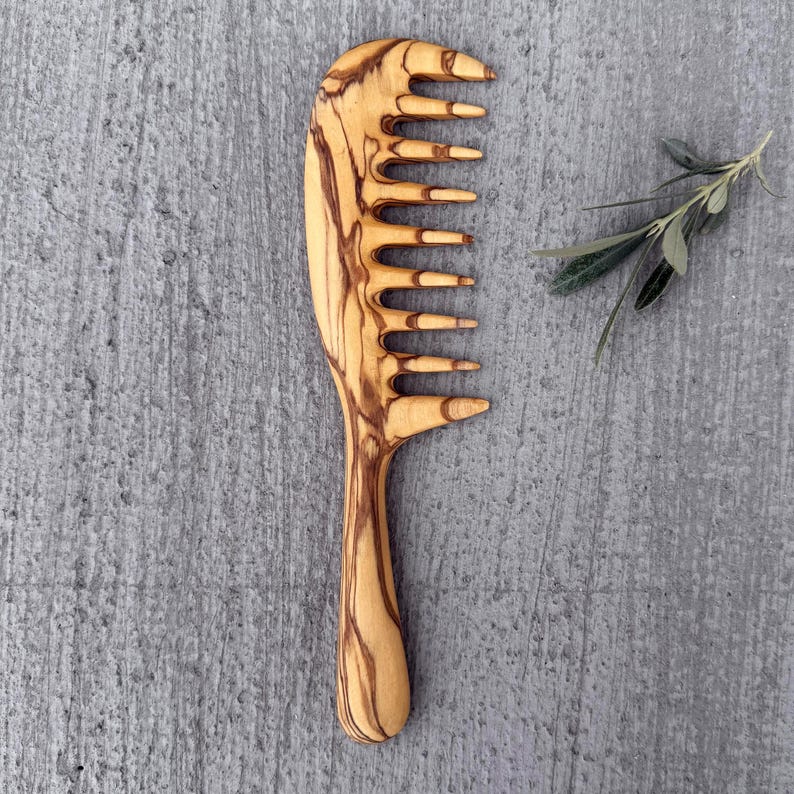 May include: A wooden comb with a natural wood grain pattern. The comb has a wide handle and a row of teeth on one side. The comb is made of olive wood.