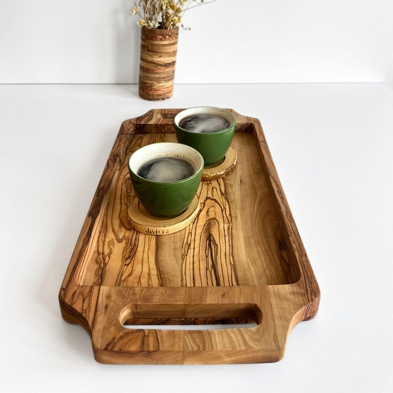 May include: A wooden serving tray with two green ceramic mugs filled with coffee, each resting on a wooden coaster. The tray has two handles and a natural wood grain pattern.