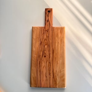 May include: A rectangular wooden chopping board with a handle. The board is made of light brown wood with dark brown grain patterns. The handle has a hole at the top. The board is approximately 30 cm long and 15 cm wide.