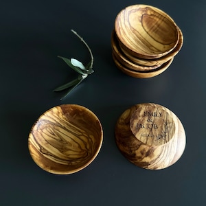May include: Four small olive wood bowls, one with the text "Emily & Jacob 12.22.2023" engraved on the bottom. The bowls are stacked on a dark gray surface.