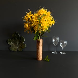 May include: A wooden vase holds a vibrant bouquet of yellow flowers with green leaves. Two empty wine glasses stand nearby. A green leaf-shaped ceramic dish is on the left. The scene is set against a dark background.