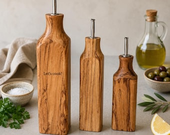 May include: Three olive wood oil dispensers of varying heights, each featuring a silver metal pour spout. The tallest dispenser is engraved with the text Let's cook! The wood grain is visible on all three bottles, which have a square base.