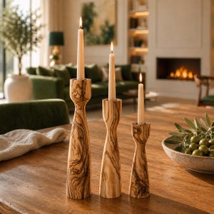 May include: Three olive wood candle holders with lit candles on a wooden table. The candle holders have a distinctive grain pattern. The candles are white and slender. A bowl of olives is in the foreground.